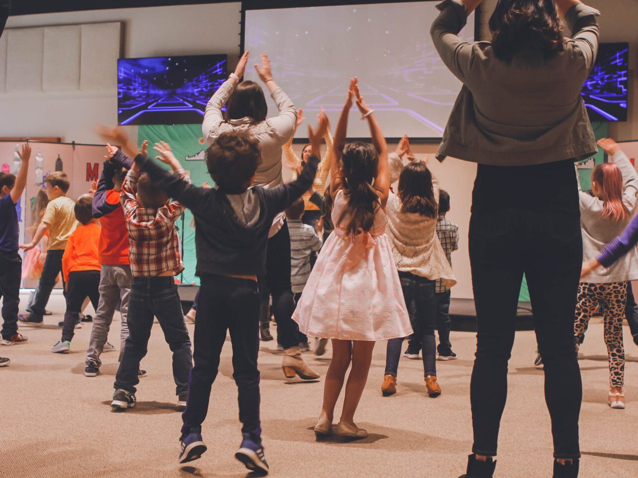 Viele kleinere Schüler, die gemeinsam und zu gleich einen Tanz präsentieren. Alle Schüler und zwei Erwachsene sind mit dem Rücken zu sehen