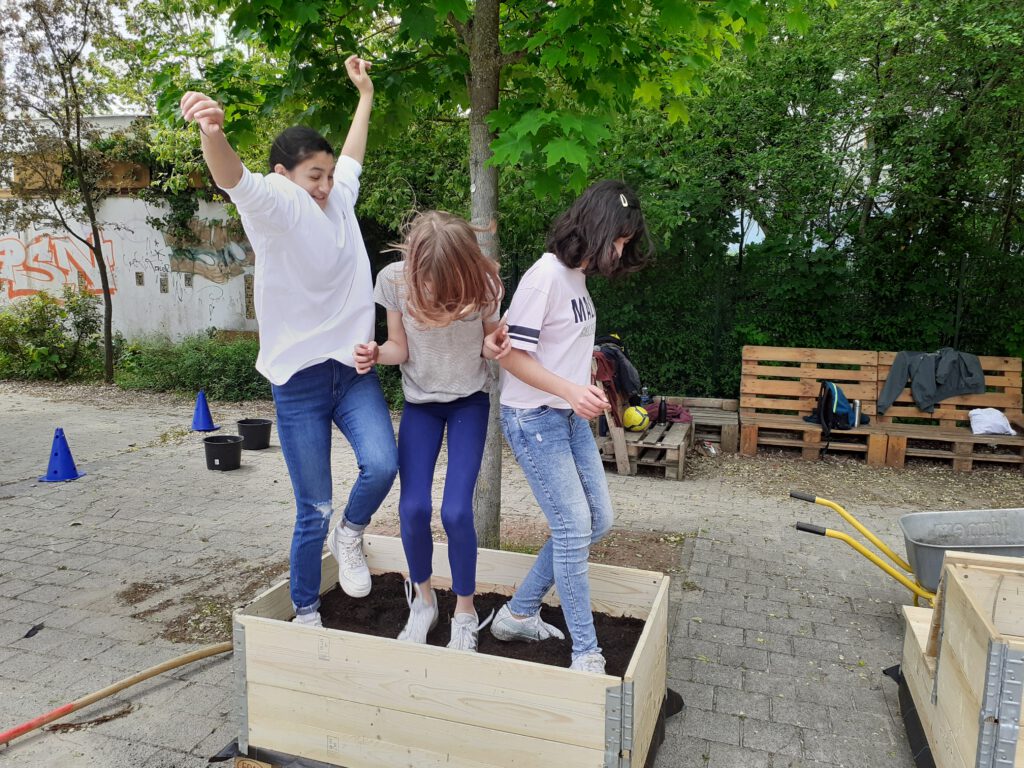 Drei Mädchen stampfen in einem Hochbeet springend Erde hernieder.