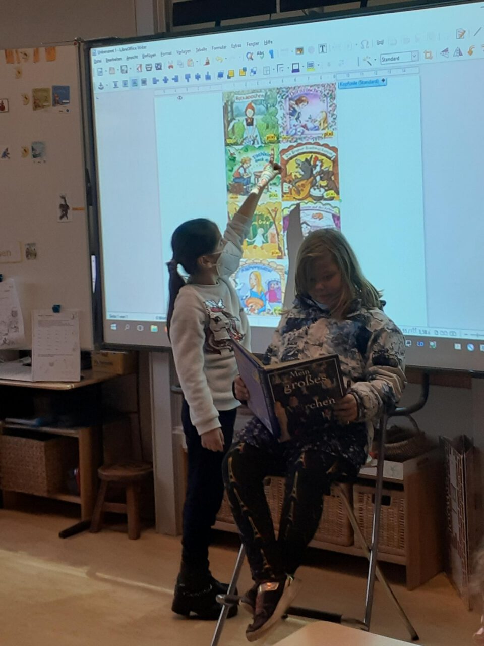 Ein Kind sitzt vor einem Whiteboard und liest vor, ein weiteres Kind steht daneben und zeigt auf Bilder