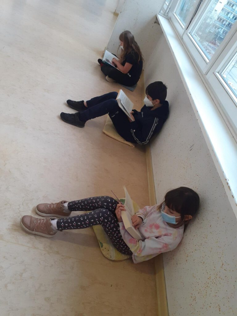 3 Kinder sitzen auf dem Boden, haben jeweils ein Buch in der Hand und lesen