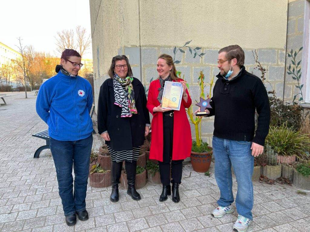 Die Schulleiterin und die koordinierende Erzieherin nehmen die Urkunde vor dem Haortgebäude von zwei Mitarbeitern des PSV Olympia entgegen.