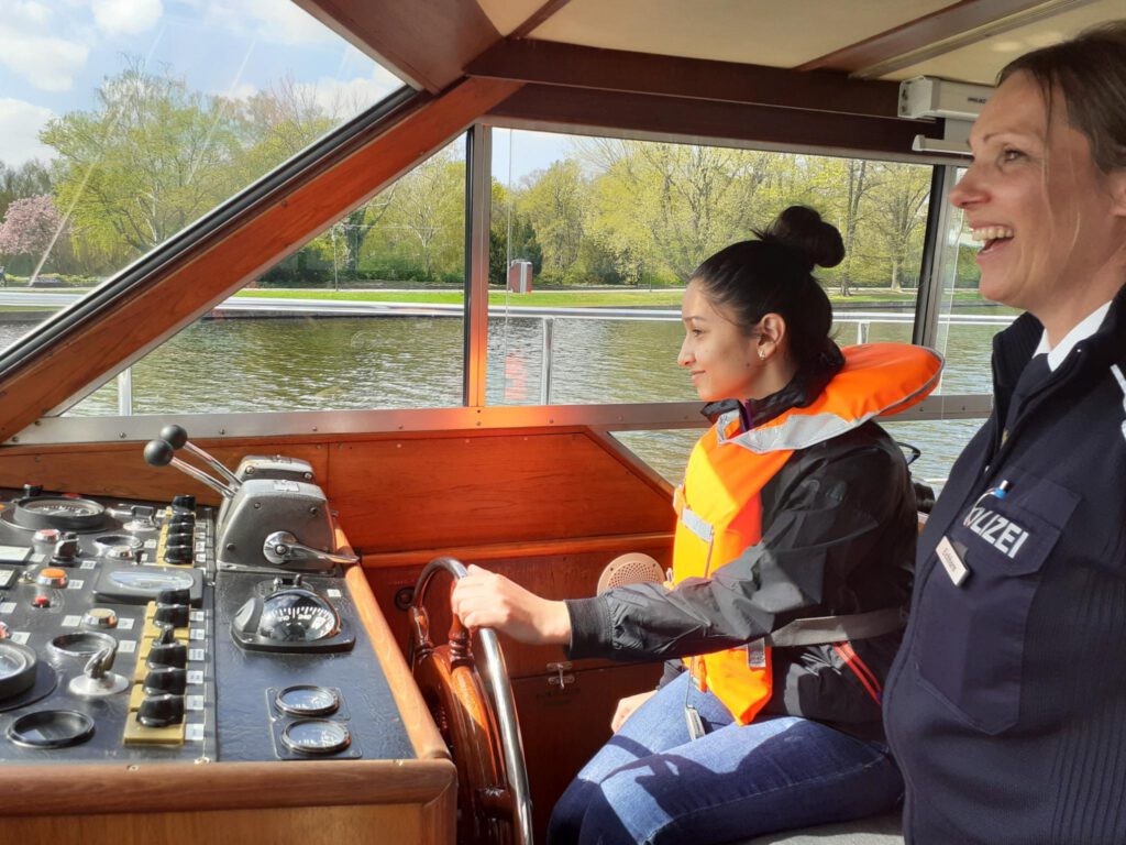 Mädchen sitzt am Steuer eines Polizeibootes, eine Polizistin steht neben ihr