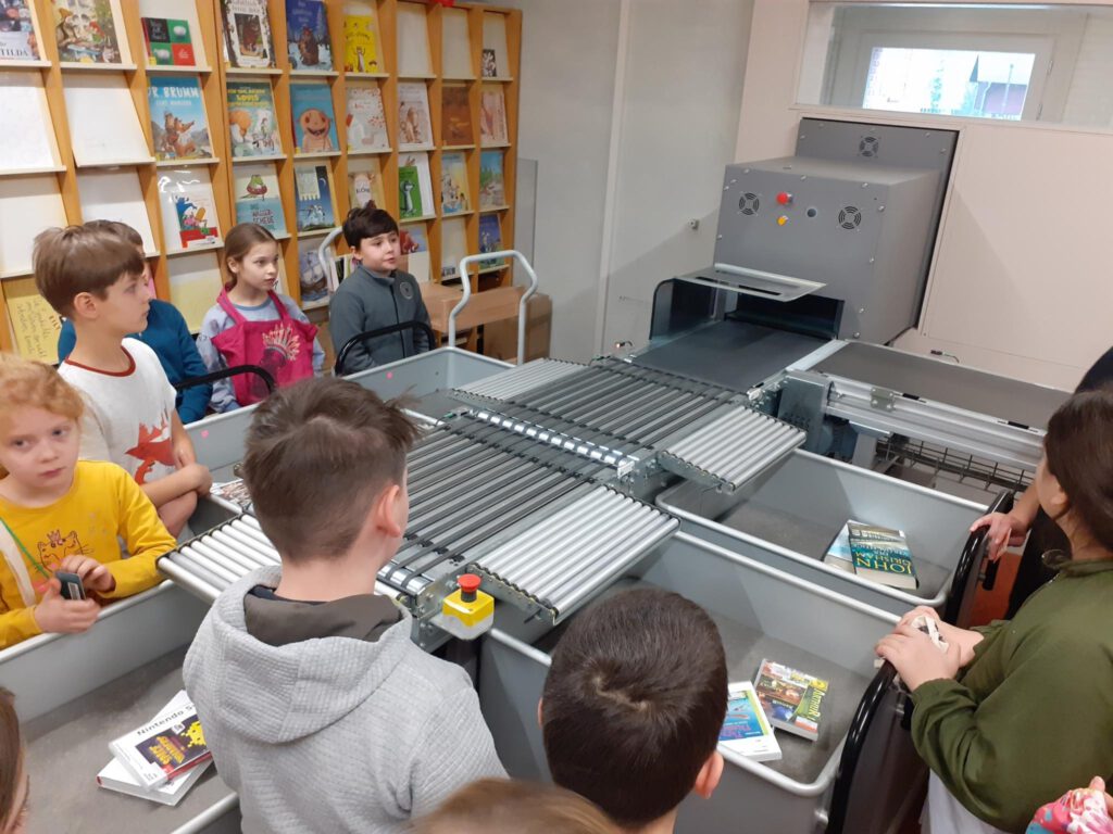 Kinder stehen um eine Maschine welche Bücher sortiert